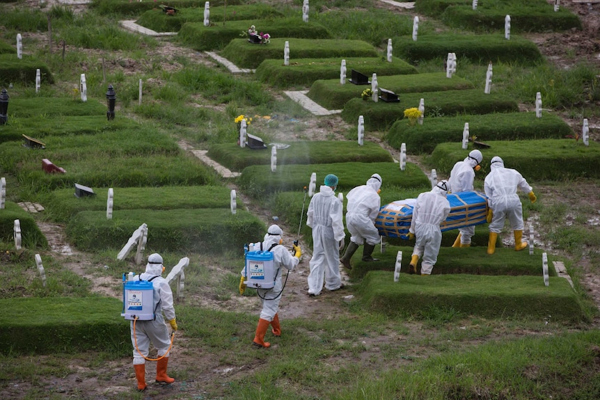 A group of men in full PPE carry a coffin wrapped in tarps through a cemetery