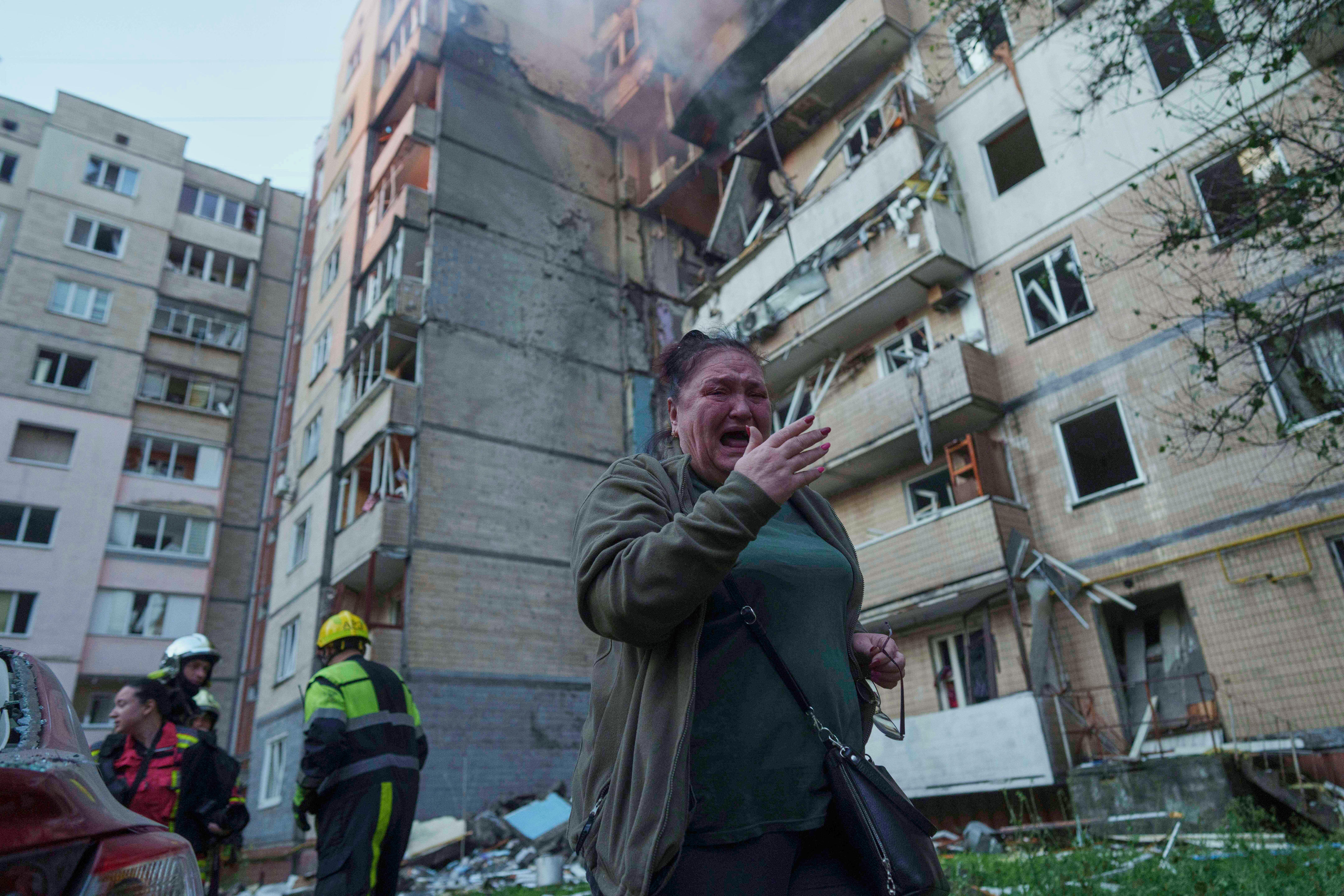 A Ukrainian woman wearing a green top and jacket crying and standing in front of a bomb-destroyed smoking building