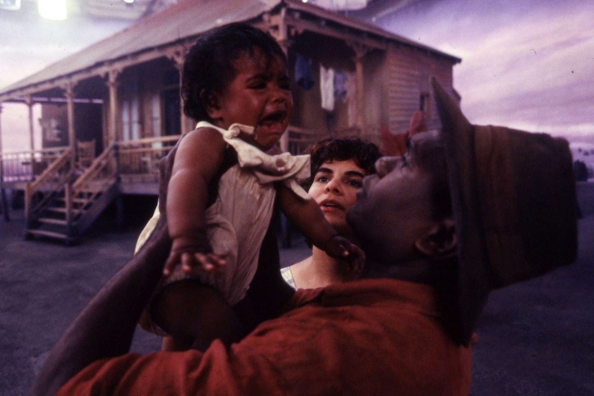 A man holds a crying baby, a woman looks on. Country house in background.