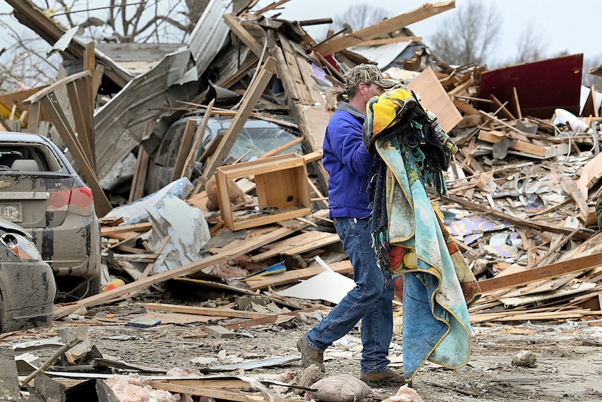 A woman carries items in her arms as she walks past a house that has been stroyed.