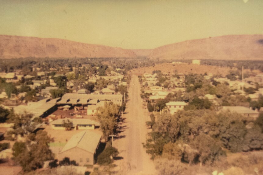 A faded photo, taken from a height, showing a long road with low buildings on either side, running towards some ranges.