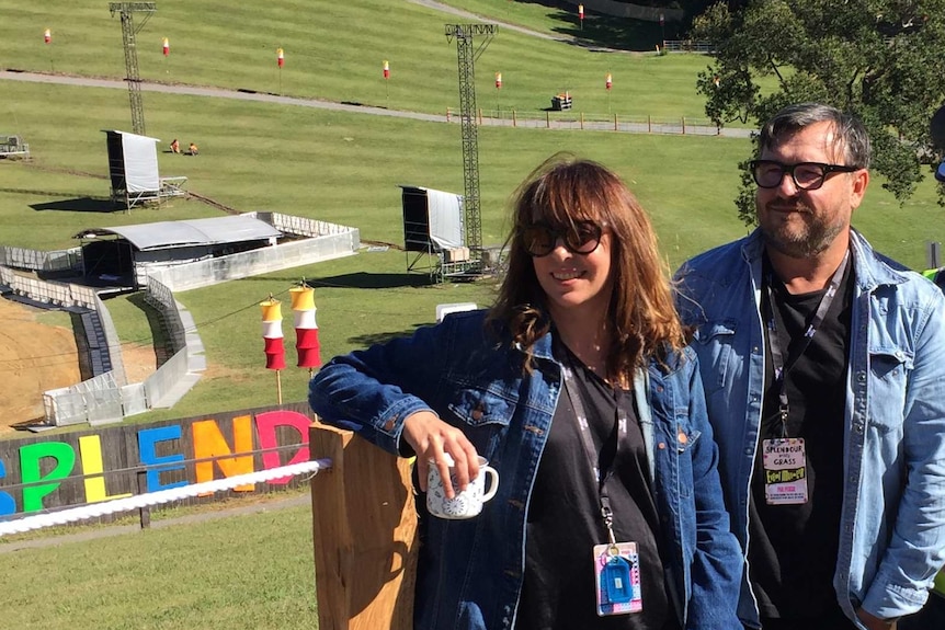 A smiling woman and a man, both wearing denim jackets, stand in front of a festival ground.