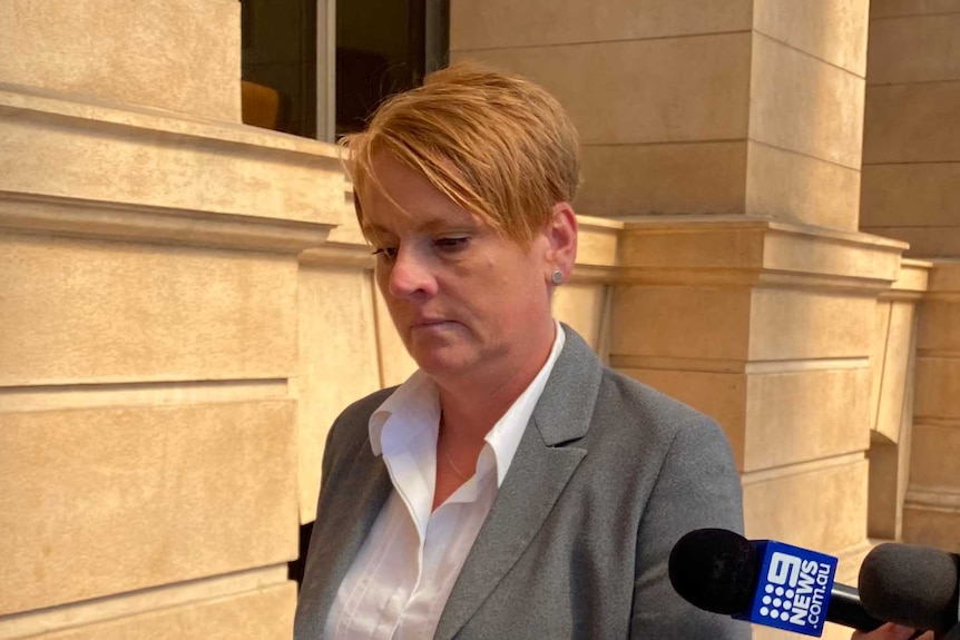 A woman with short hair wearing a grey jacket being trailed by reporters outside a court building