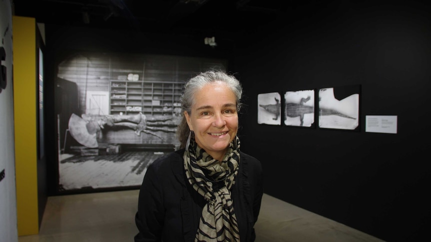 Vanessa Finney wears a black shirt and black and grey scarf. She smiles against a backdrop of photographs.