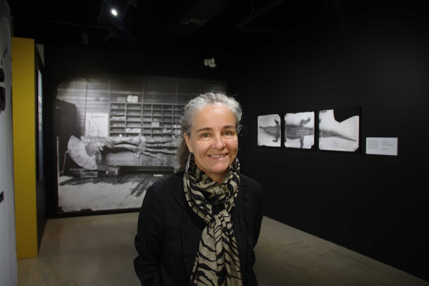 Vanessa Finney wears a black shirt and black and grey scarf. She smiles against a backdrop of photographs.