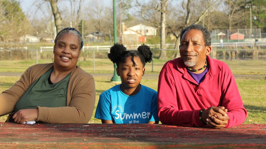 A man, child and woman sit a picnic bench in a park-like setting 