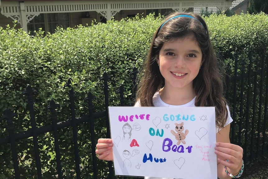 A young girl stands outside holding a hand made sign that says we're going on a bear hunt.