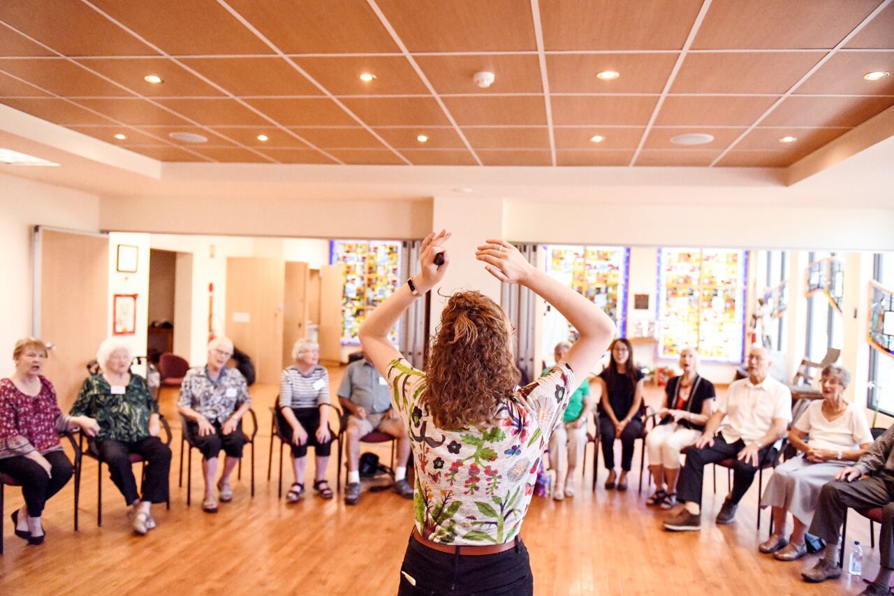 A view from behind someone conducting with arms raised a group of people singing while sitting on chairs in a semi-circle.