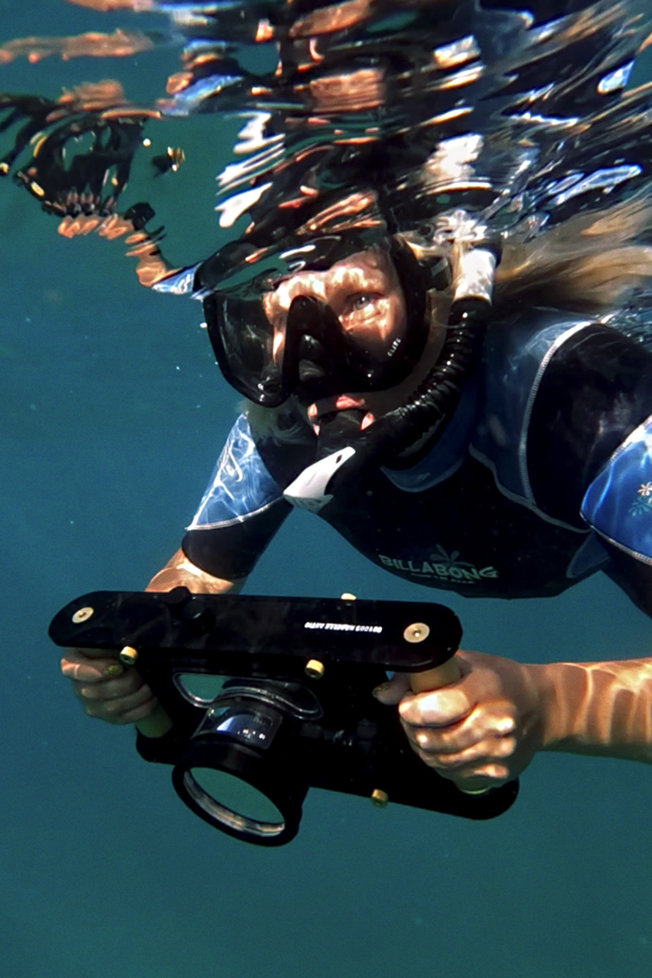 Close-up of photographer Narelle Autio underwater wearing a wetsuit, mask and snorkle, and holding a large underwater camera 