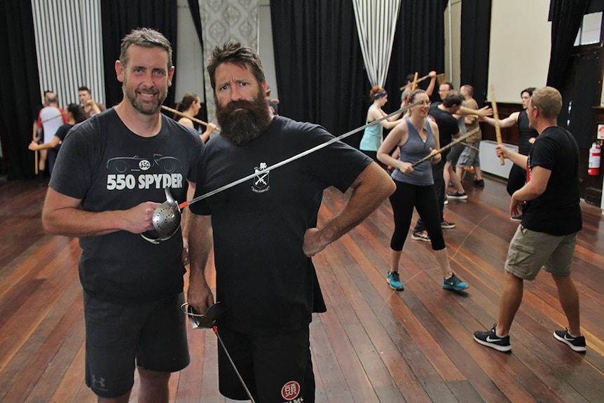 Two men wielding swords pose in front of a room full of actors who are training with bamboo poles.