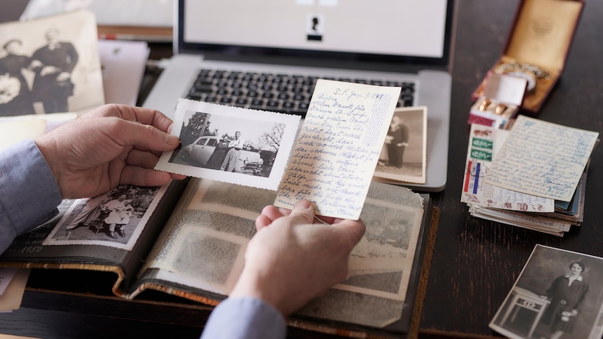 A man's hands holding an old photo and letter, with laptop and photo album in the background