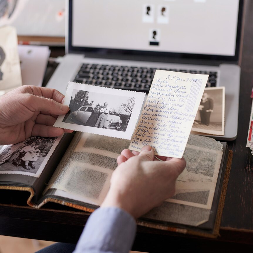 A man's hands holding an old photo and letter, with laptop and photo album in the background