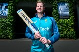Steve Smith smiles while holding his cricket bat in front of a leafy backdrop