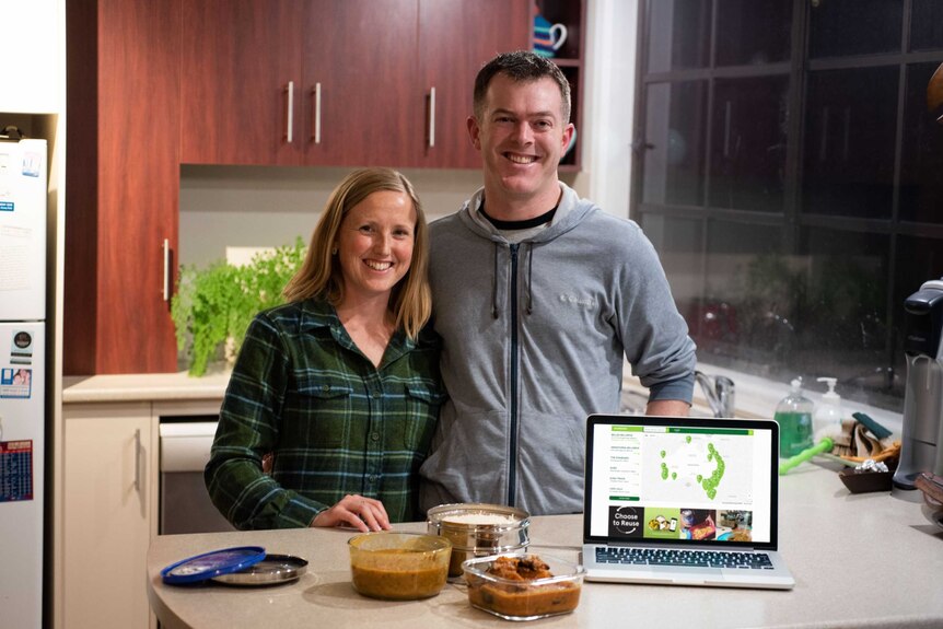 A woman and man smiling and standing near a kitchen bench with the trashless takeaway website on a laptop