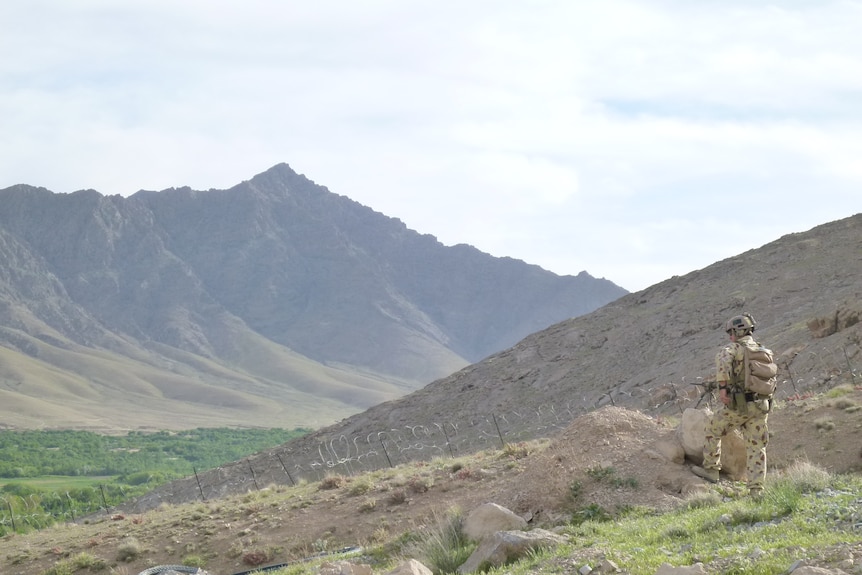 an ADF soldier in uniform stands guard near a barbed wire fence among hills in Afghanistan