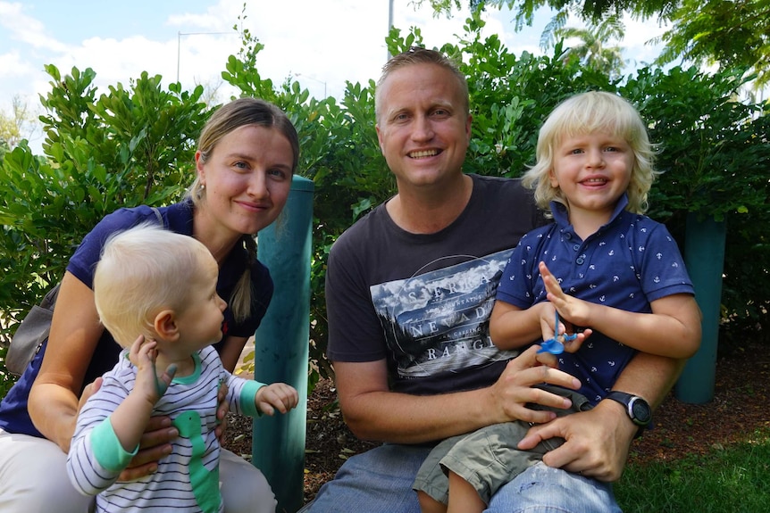 Matthew Kerle and his wife hold their two young boys. They are all smiling at each other and are outside in the sunshine.