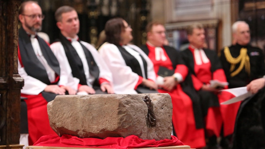 A large grey stone with a bolt is sitting on red velvet fabric. In the background are religious figures.