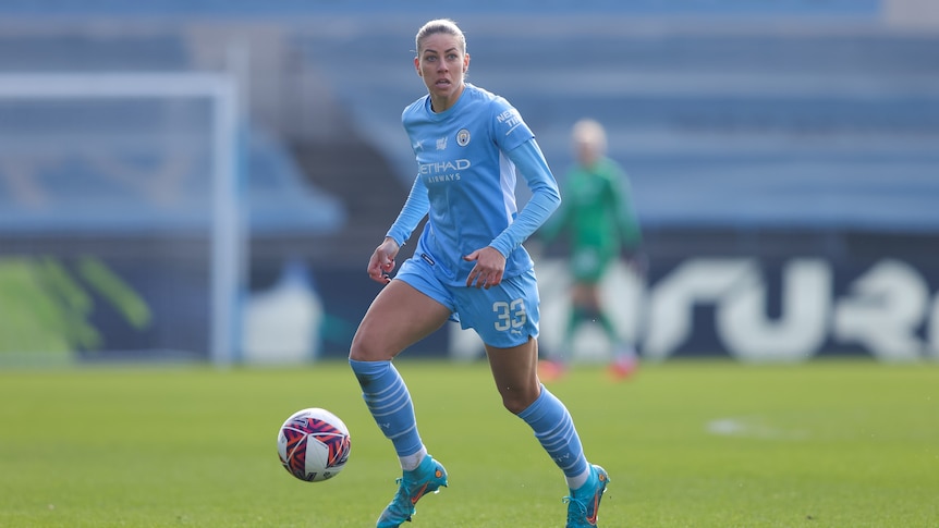 A female soccer player wearing light blue looks up with the ball at her feet during a match