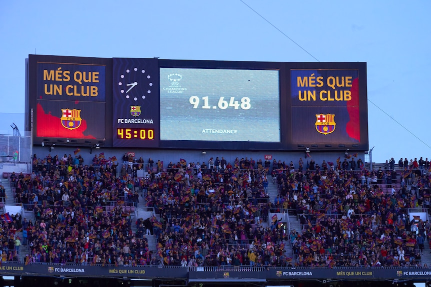 A giant LED screen on top of a grandstand with the FC Barcelona logo displays the time and a sign "91,648 attendance".