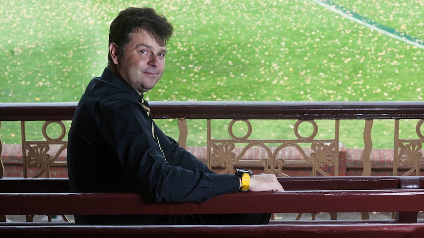 A man with short, brown hair sitting in a grandstand looking back at the camera, with the green field in the background.