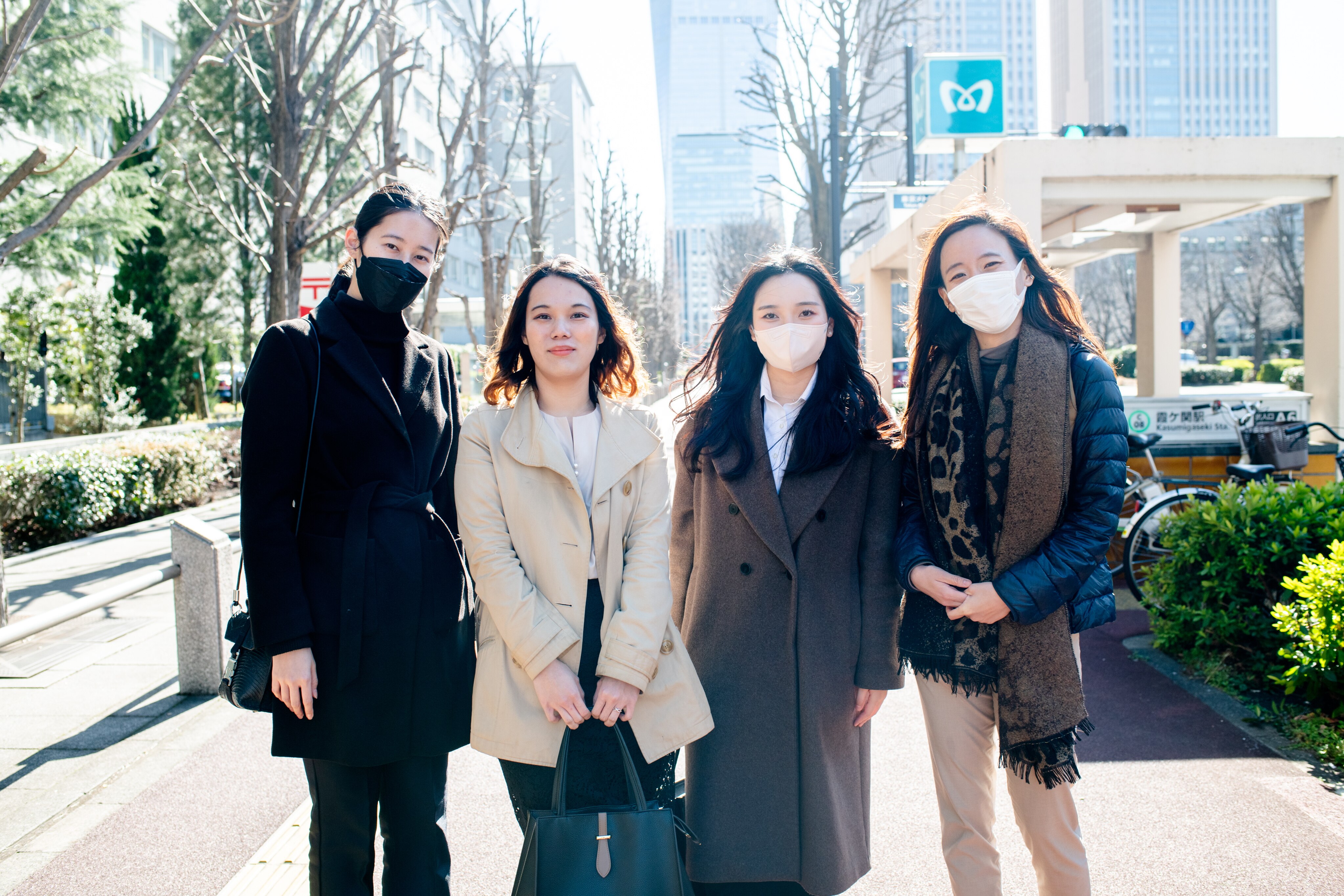 Four women stand in coats and winter jackets outside on a sunny day in a city setting. Three are wearing facemasks