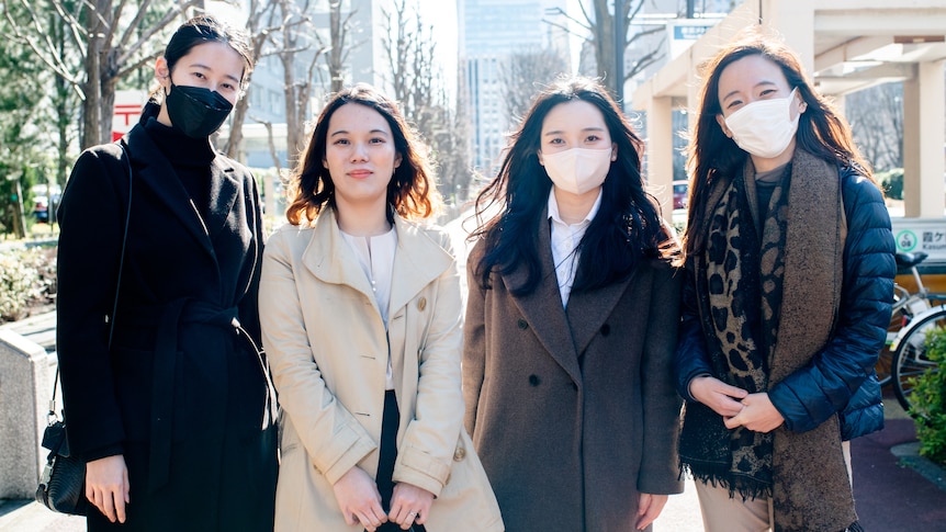 Four women stand in coats and winter jackets outside on a sunny day in a city setting. Three are wearing facemasks