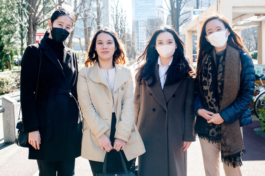 Four women stand in coats and winter jackets outside on a sunny day in a city setting. Three are wearing facemasks