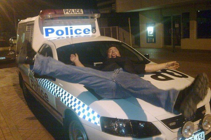 Damian Maxfield lies on a parked police car with his legs and arms spread, tongue stuck out.