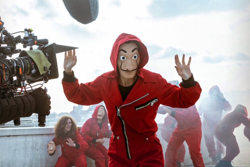 A person in a red jump suit and mask puts their hands up with a film camera in shot