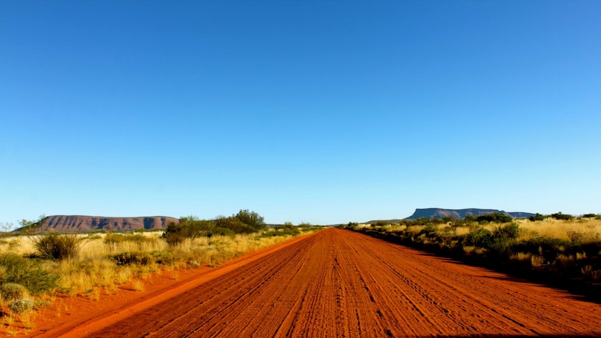 A straight red dirt road leading to Kintore. Two large rocky mountains are located in the background on either side of the road.