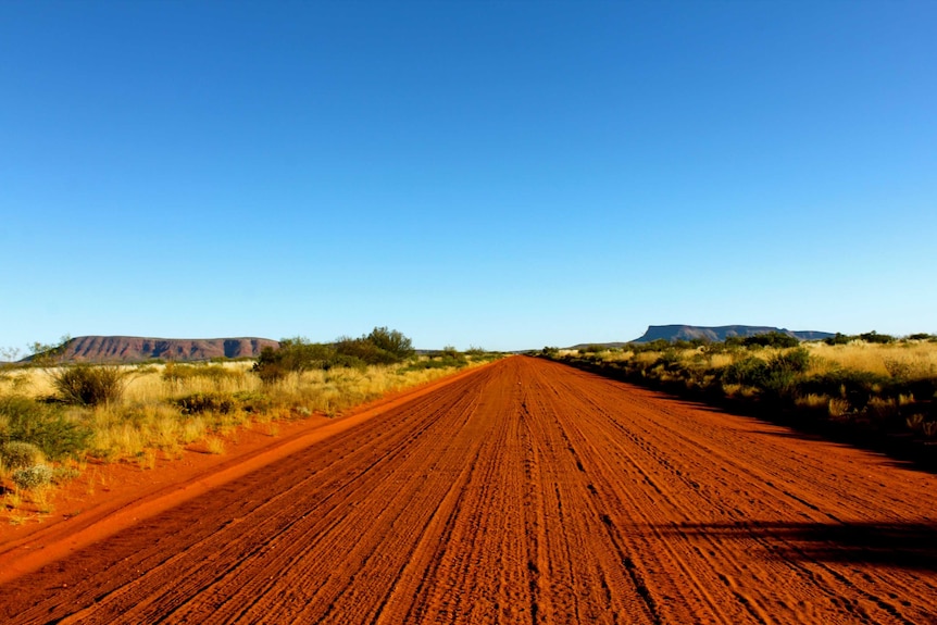 A straight red dirt road leading to Kintore. Two large rocky mountains are located in the background on either side of the road.