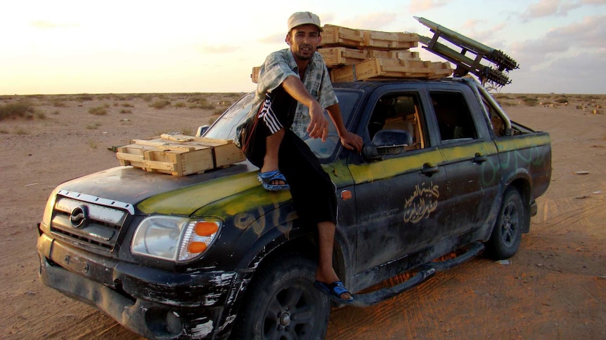 A man sits on the bonnet of a truck loaded with weapons.