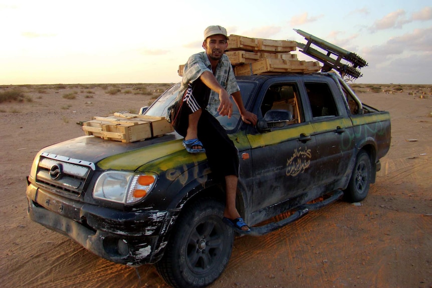 A man sits on the bonnet of a truck loaded with weapons.