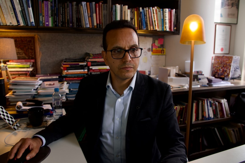  A serious man in dark suit jacket, light open shirt, wears glasses, hand on mouse, looks away from camera, books, lamp behind.
