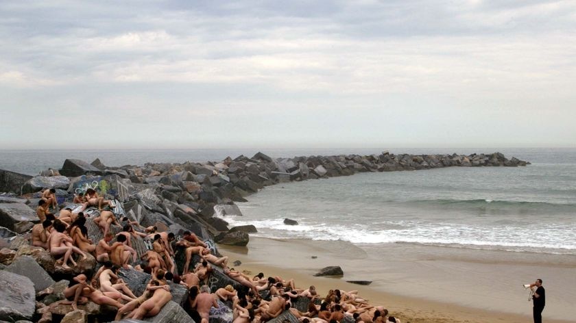 Naked models lie on sand and rocks by a beach.