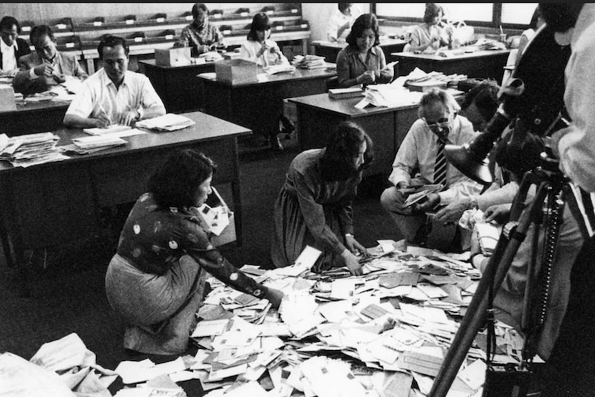 Black and white photo of people sorting a pile of letters on the floor and others sitting at desks opening letters.