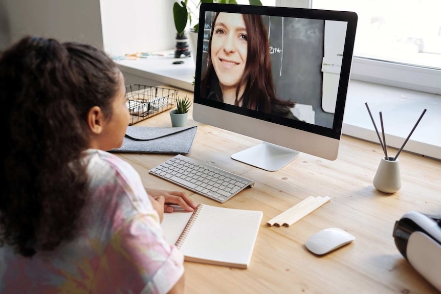 A girl sits in front of a computer, which shows a teacher delivering an online lesson.