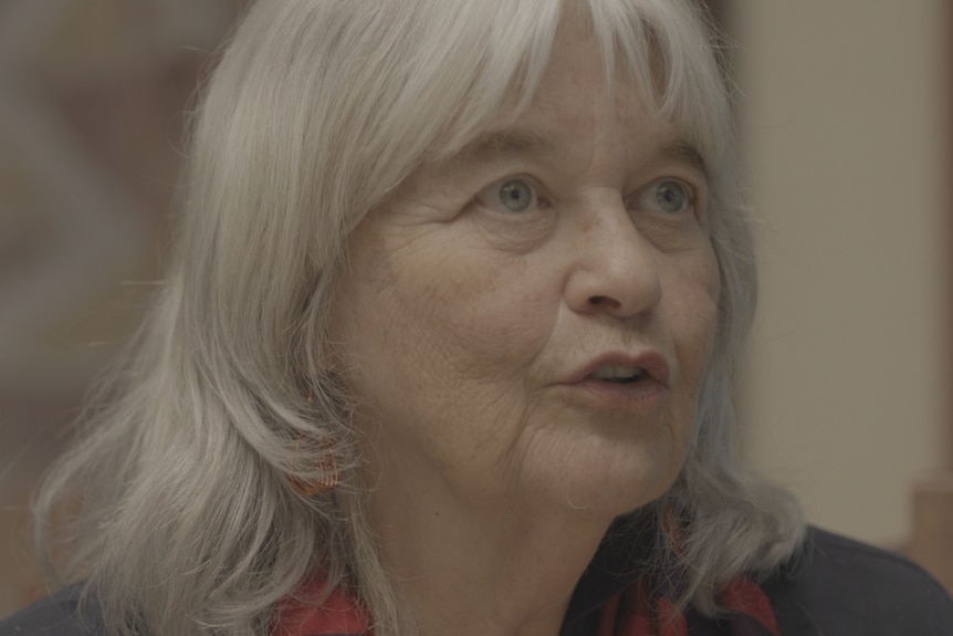 A woman with grey hair and a brown shirt talks with someone.