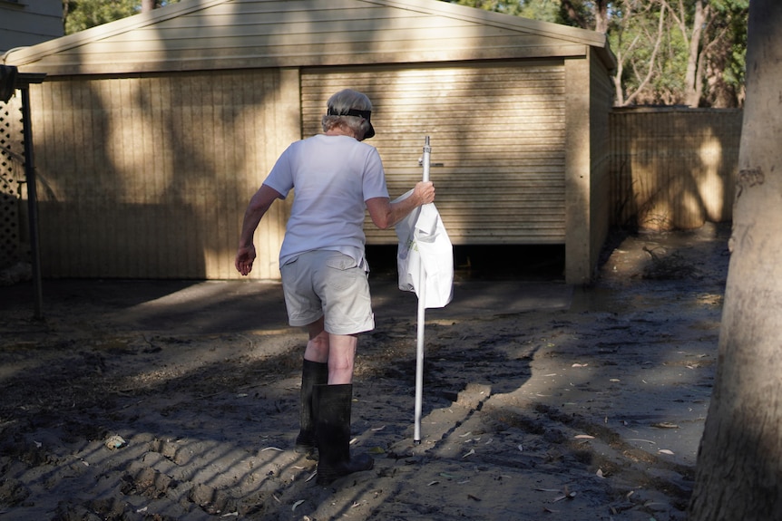A woman walks away from the camera in a muddy driveway while holding a pole and a bag