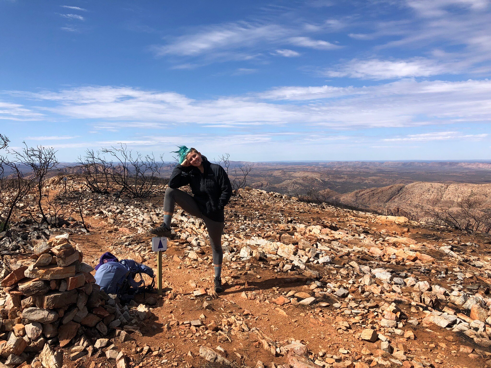 A person stands with one foot on a signpost upon a rocky dirt track with blue sky behind them.