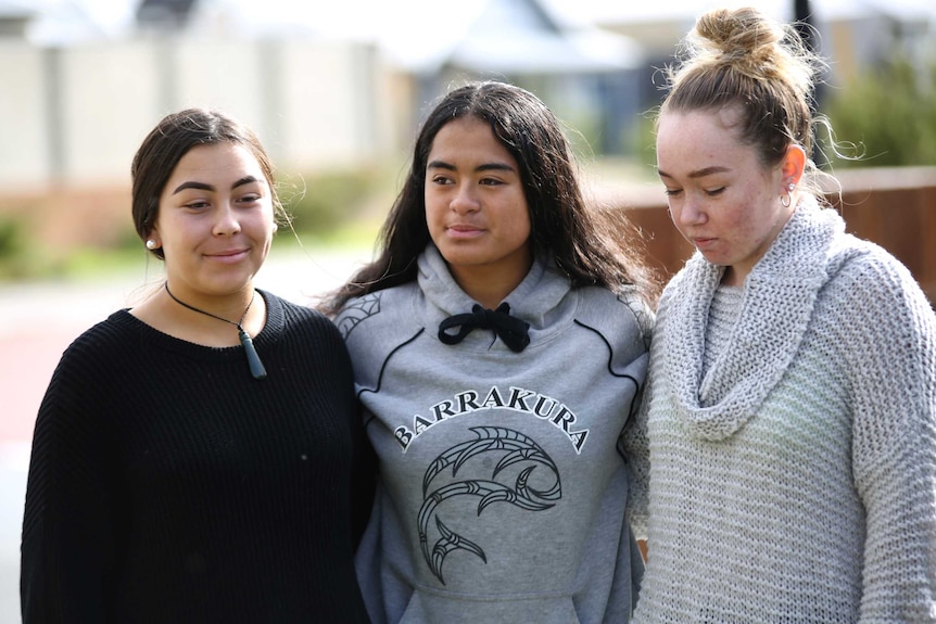 Three teenage girls in jumpers stand with arms around each other on a suburban street.