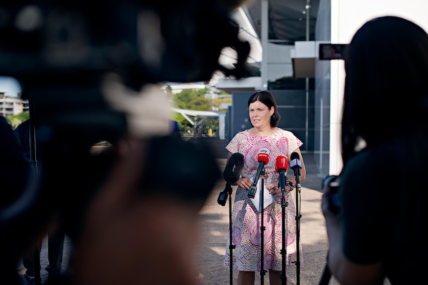 A woman with black hair standing at several microphones