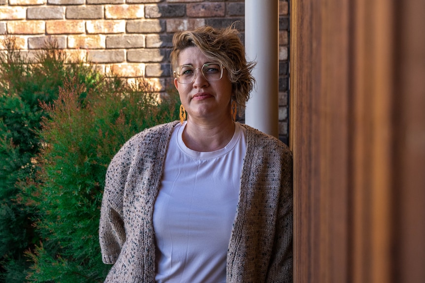 Larissa is looking at the camera leaning against a wooden wall. She is wearing a white t-shirt with a brown cardigan