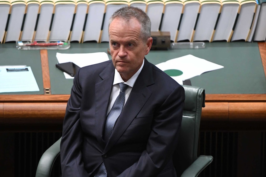 Bill Shorten wears a navy suit and grey tie as he sits on green chair with green and wood panelled desk behind him.