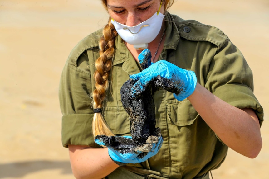 A woman with a long plait wearing army fatigues, blue gloves and a mask looks at a glob of tar