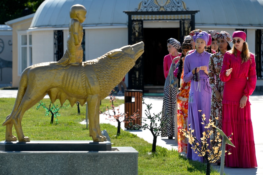 Models dressed in sparkly clothing line up next to a gold statue. 