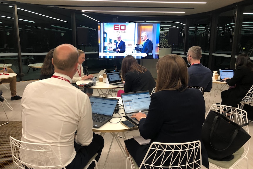 Journalists in front of laptops watching TV screen of two leaders standing behind lecturns debating.