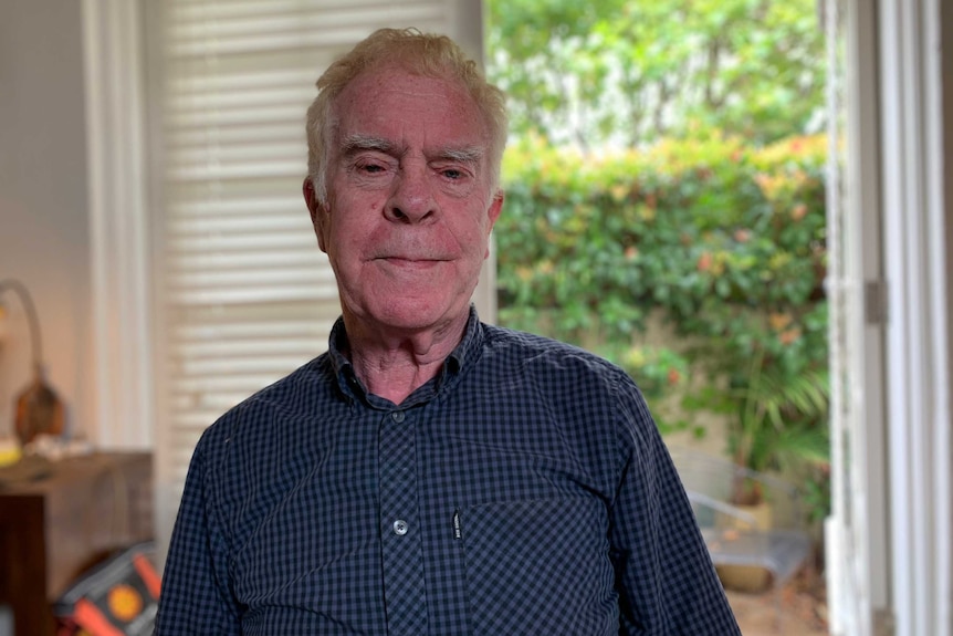 An elderly man in a blue and black checkered shirt, with a door open to the garden behind him.