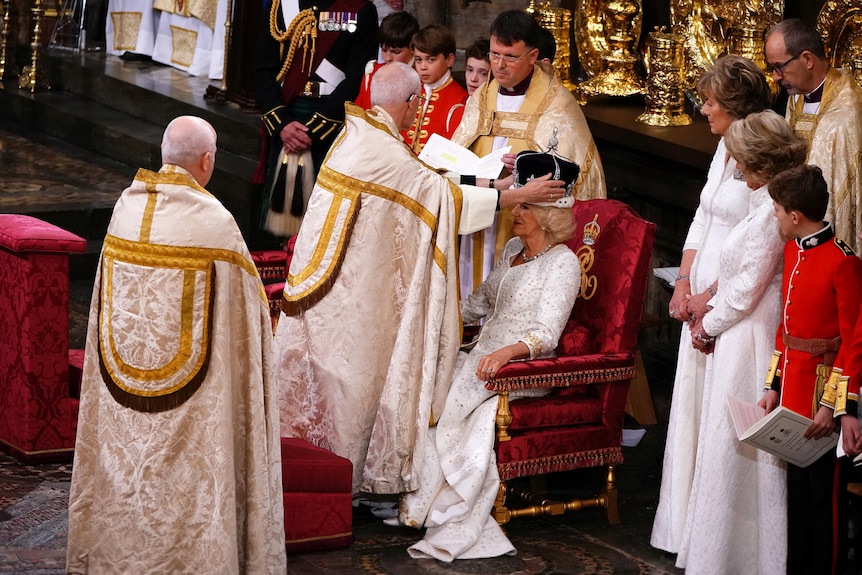 A man in a robe placing a crown on Camilla's head while she sits in a red chair. 