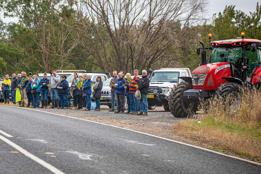 Mourners holding yellow flowers stand by the highway, with a red tractor parked alongside.